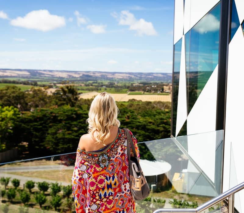 Rear view of a blonde woman wearing a vibrant, colorful patterned dress looking out from a high glass balcony over distant agricultural fields.