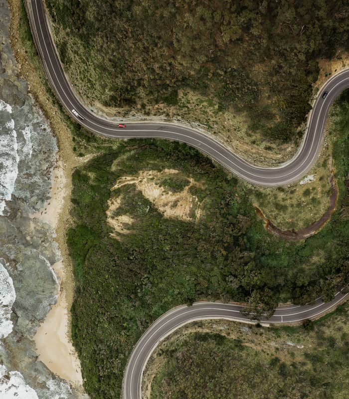 Looking straight down from a drone at the dark grey asphalt of the Great Ocean Road as it winds in tight loops between a dense green forest and a white-water surf beach.