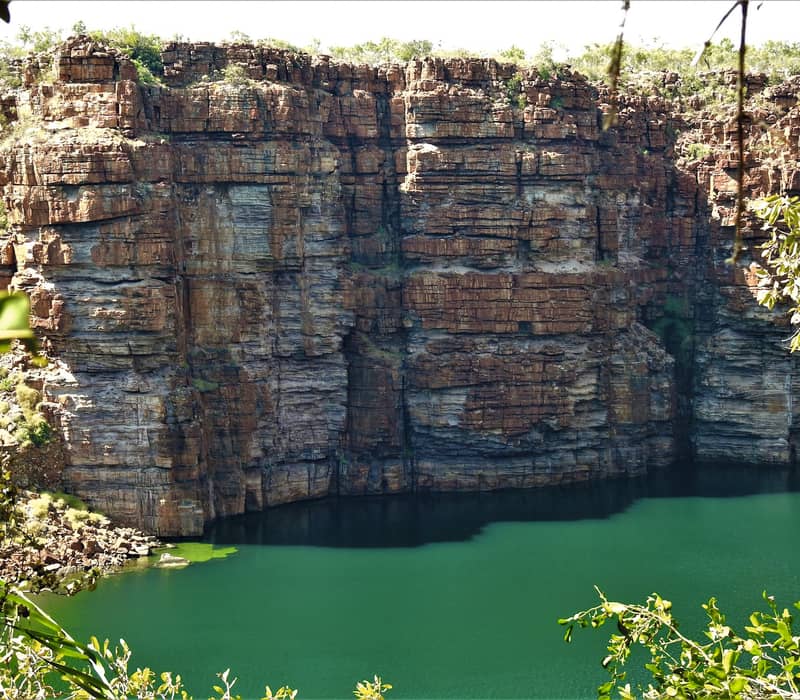 Close-up of sheer red and orange sandstone cliff faces meeting a calm green river in the Kimberley.