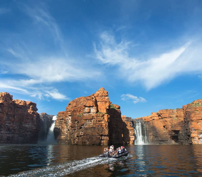A small boat on a dark river approaching two tall waterfalls cascading down massive red rock cliffs under a blue sky.