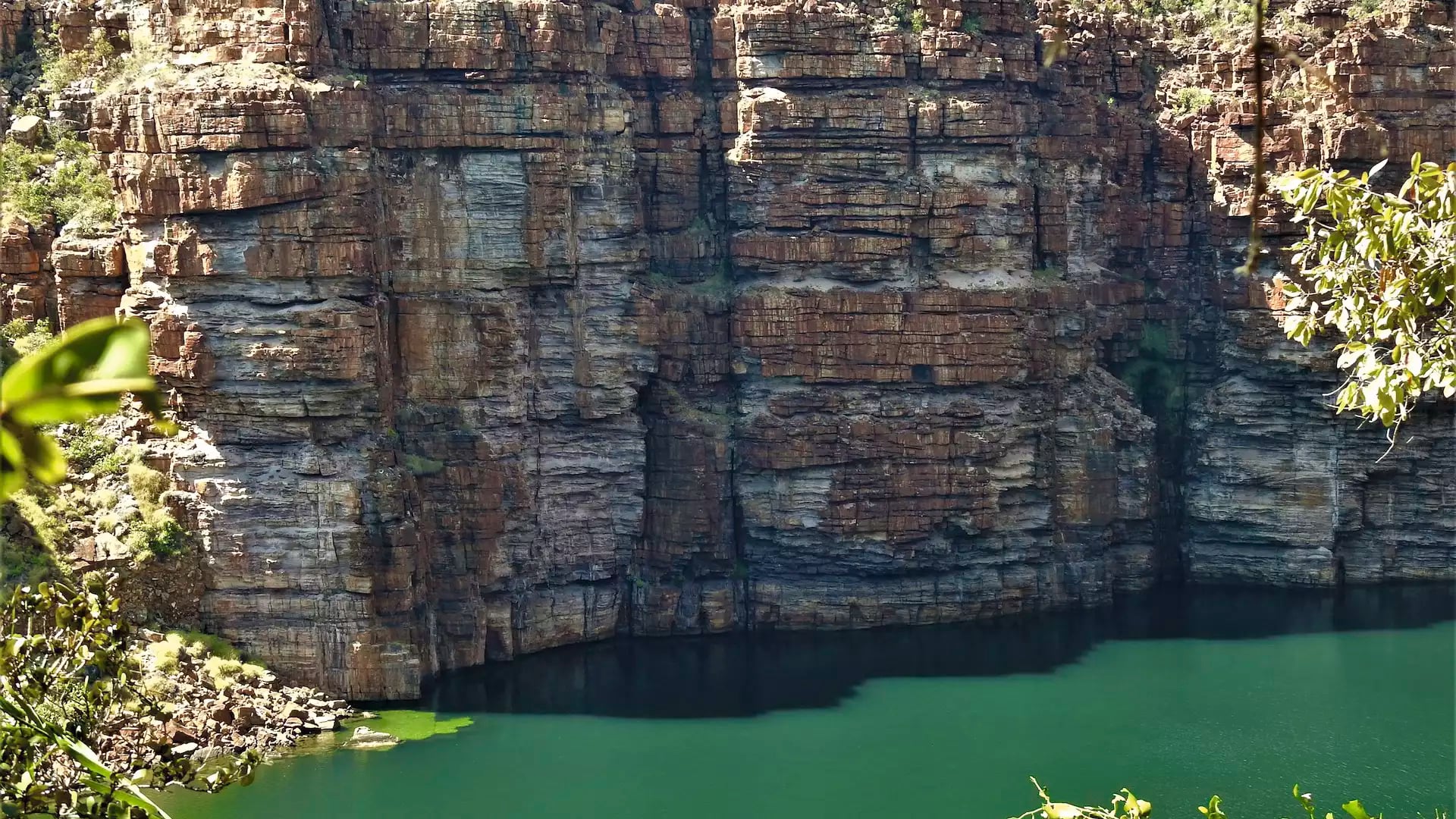 Close-up of sheer red and orange sandstone cliff faces meeting a calm green river in the Kimberley.