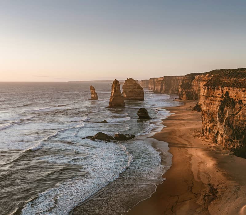A row of massive limestone pillars stands in the surf along a sandy beach and high, sheer cliff line during a soft orange sunset on the Great Ocean Road.