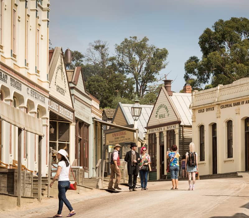 A street scene at Sovereign Hill shows historical buildings including an Apothecaries Hall and a Colonial Bank, with people in modern and period clothing walking on the dusty street.