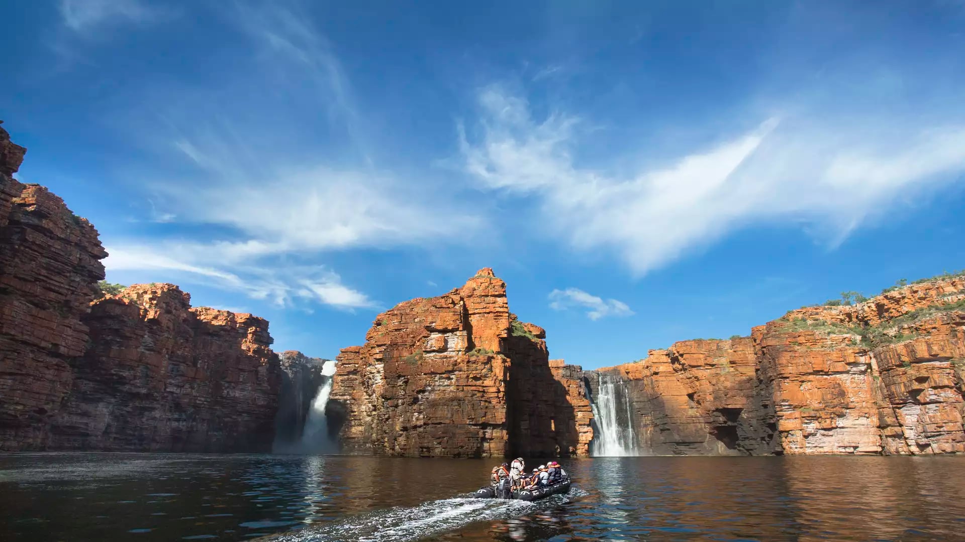 A small boat on a dark river approaching two tall waterfalls cascading down massive red rock cliffs under a blue sky.