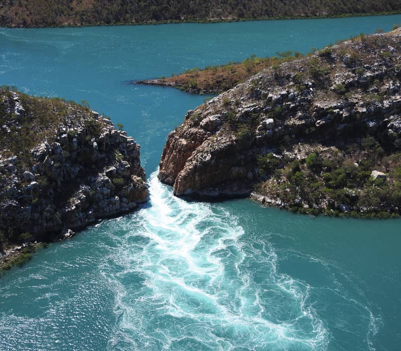Aerial view of turquoise water rushing through a narrow gap between two rocky islands in the Kimberley, creating a waterfall effect.