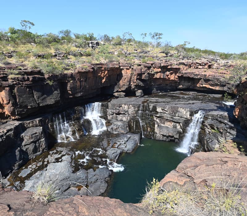 High-angle shot of a waterfall cascading over dark rocky ledges into a deep green pool in a rugged canyon.