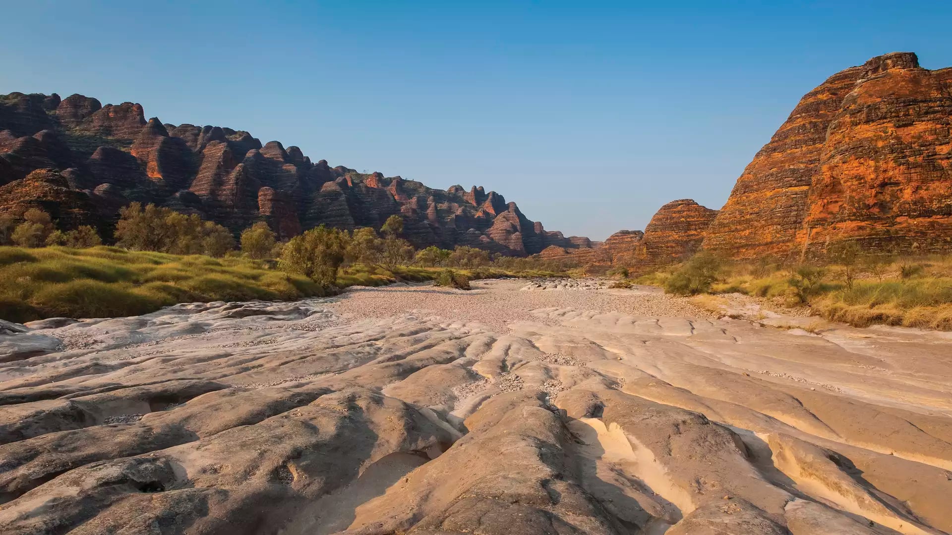 Wide landscape shot of a dry, sandy creek bed leading to the striped orange and black rock domes of the Bungle Bungles.