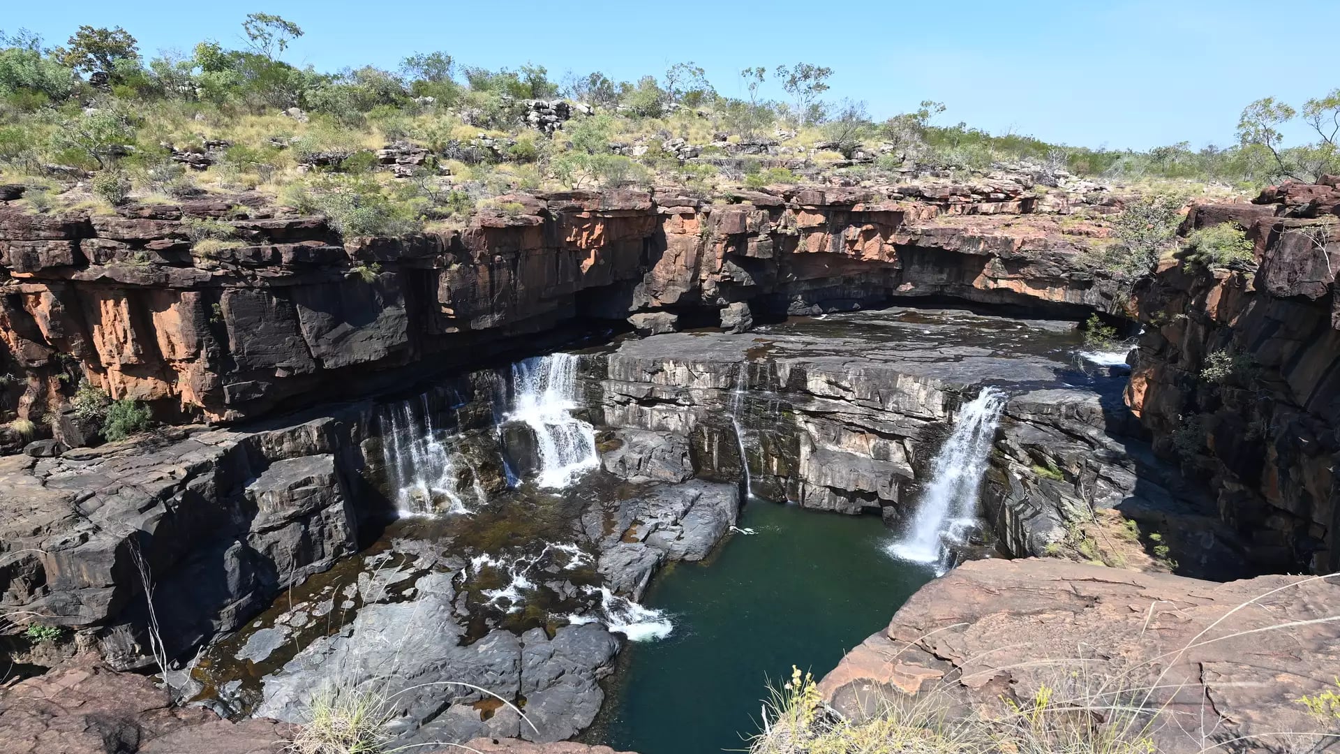 High-angle shot of a waterfall cascading over dark rocky ledges into a deep green pool in a rugged canyon.