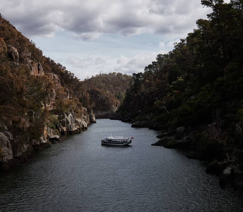 A tour boat on a dark river between steep rocky cliffs under a cloudy sky in Tasmania.