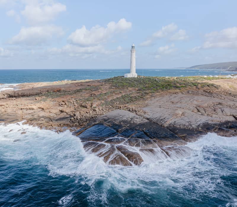 Waves crash against the dark rocks below the white Cape Leeuwin Lighthouse at the tip of Augusta in the Margaret River Wine Region.