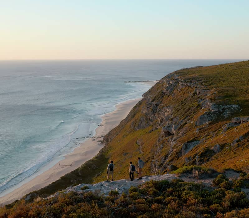Three hikers stand on a clifftop at sunset overlooking a long, white sand beach on the Cape to Cape Track in the Margaret River Wine Region.