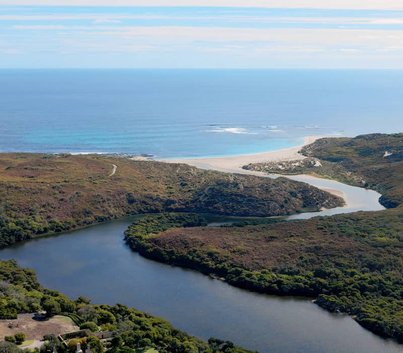 An expansive aerial shot of the Margaret River as it flows through dense green bushland to meet a white sandbar and the blue ocean at the River Mouth in Prevelly.