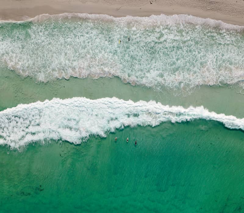 A direct overhead aerial view of surfers paddling in the emerald green water as white waves break at Gas Bay, Margaret River.