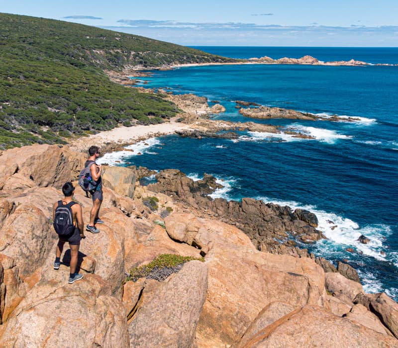 Two men stand on a high rocky cliff edge looking out over the winding coastline and turquoise bays of Contos Beach in the Margaret River region.