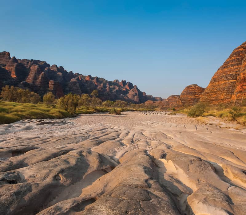 Wide landscape shot of a dry, sandy creek bed leading to the striped orange and black rock domes of the Bungle Bungles.
