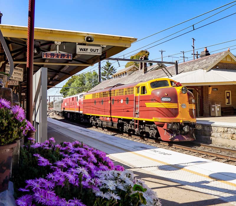 A vintage red diesel locomotive arriving at a heritage railway station platform decorated with flowers.