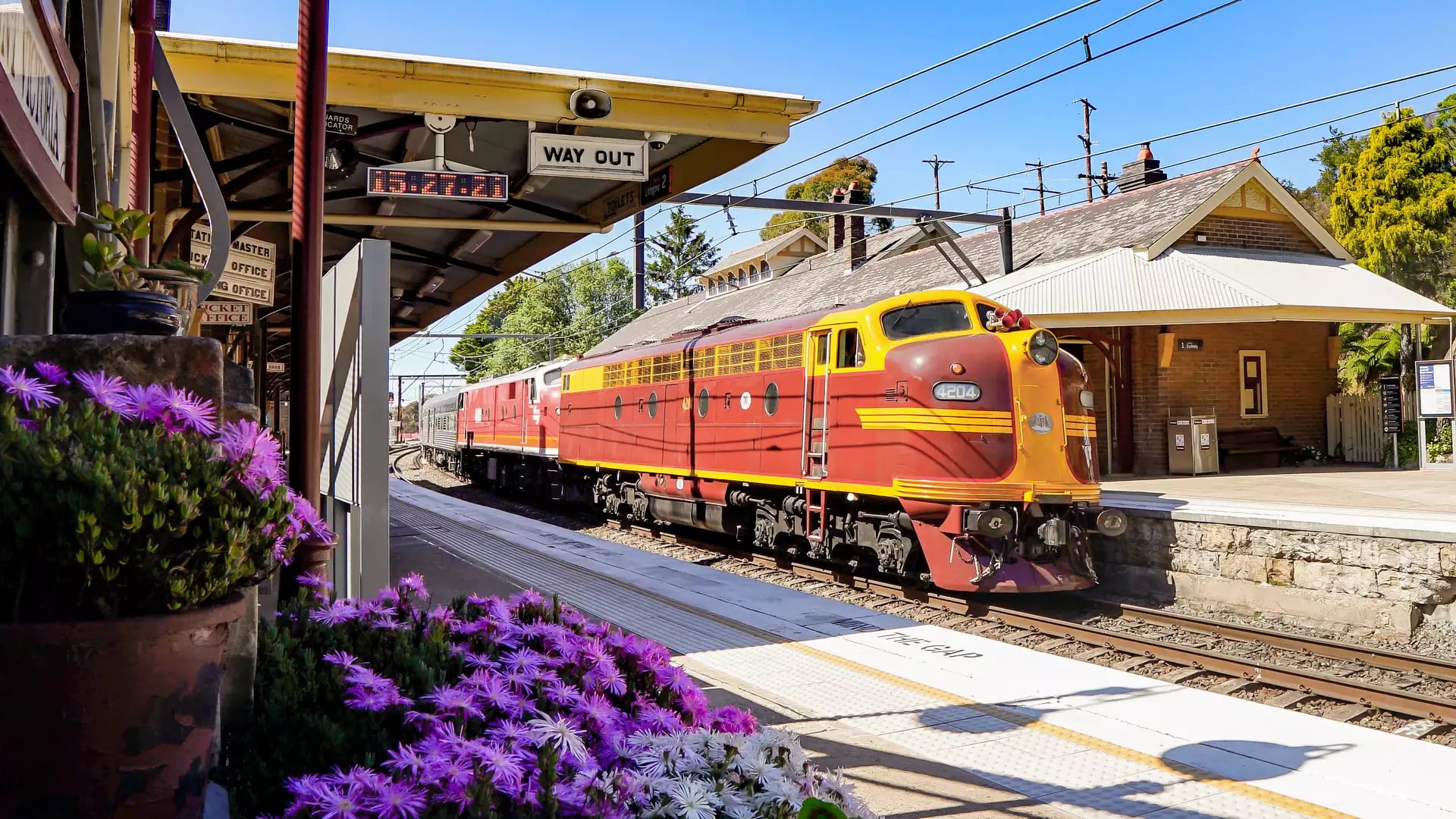 A vintage red diesel locomotive arriving at a heritage railway station platform decorated with flowers.