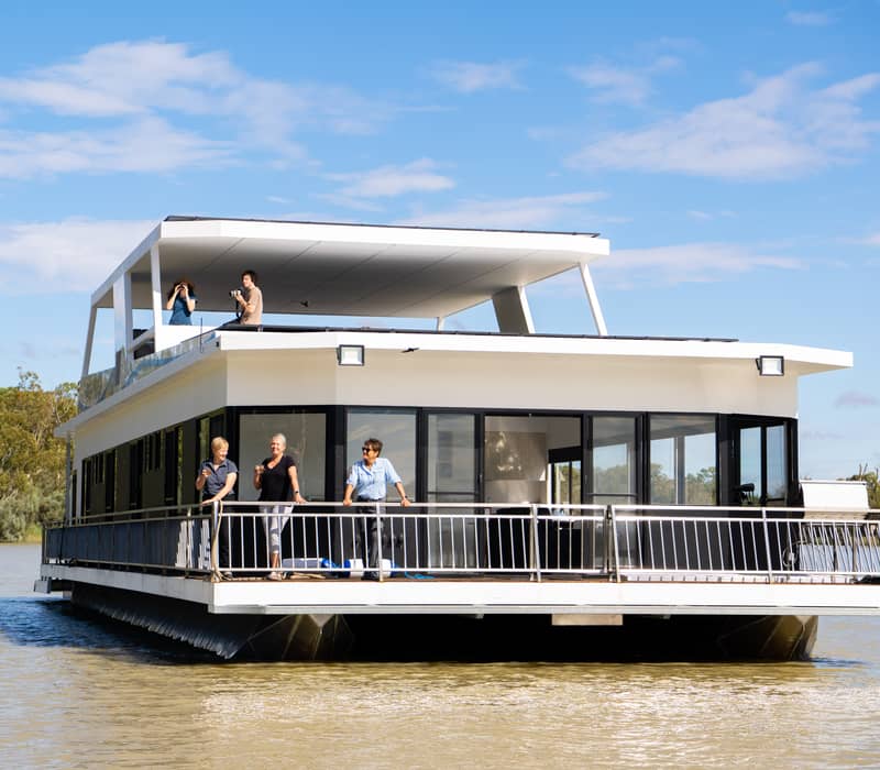 A large, modern white two-story houseboat with guests on the front and top decks cruising on the Murray River.