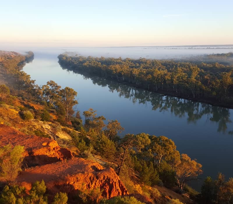 High-angle landscape view of the Murray River at sunrise with mist over the water and red orange cliffs in South Australia.