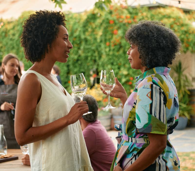 Two women holding wine glasses and talking in front of a greenery-covered winery building in South Australia.