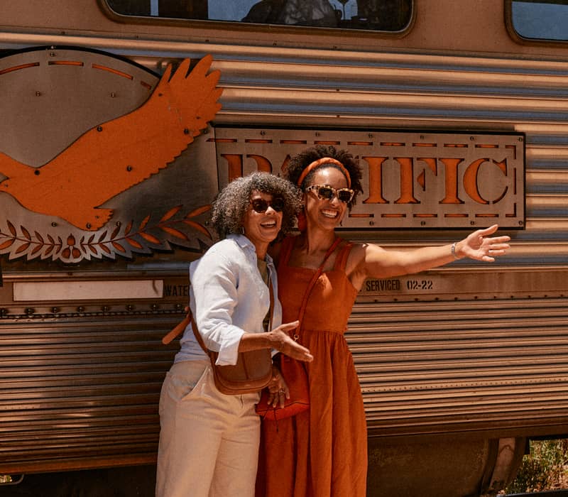 Two women smiling and waving next to the silver fluted carriages of the Indian Pacific train at a desert stop.