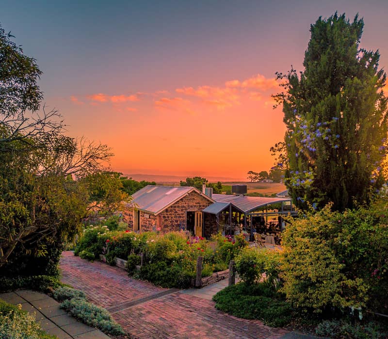 Vibrant orange and pink sunset sky over a rustic stone winery building and manicured gardens in South Australia.