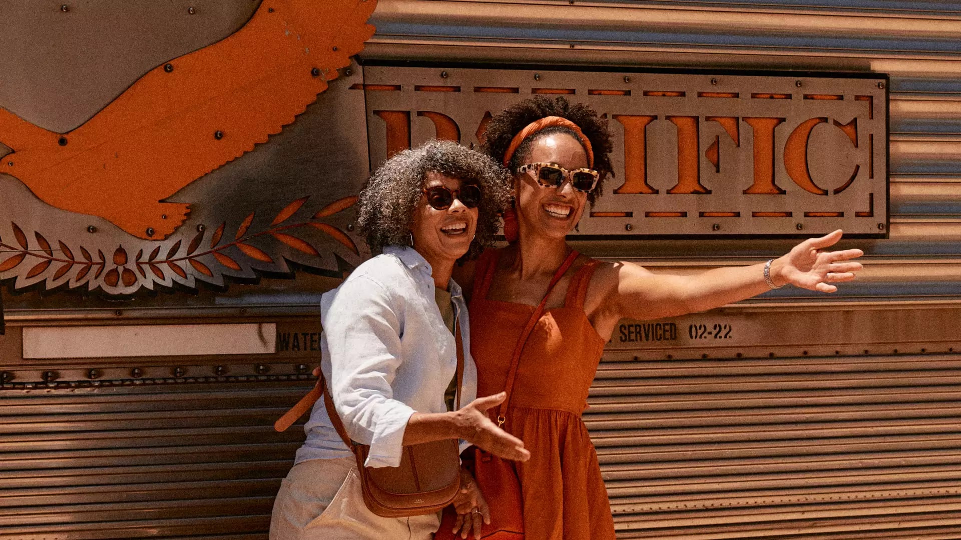 Two women smiling and waving next to the silver fluted carriages of the Indian Pacific train at a desert stop.