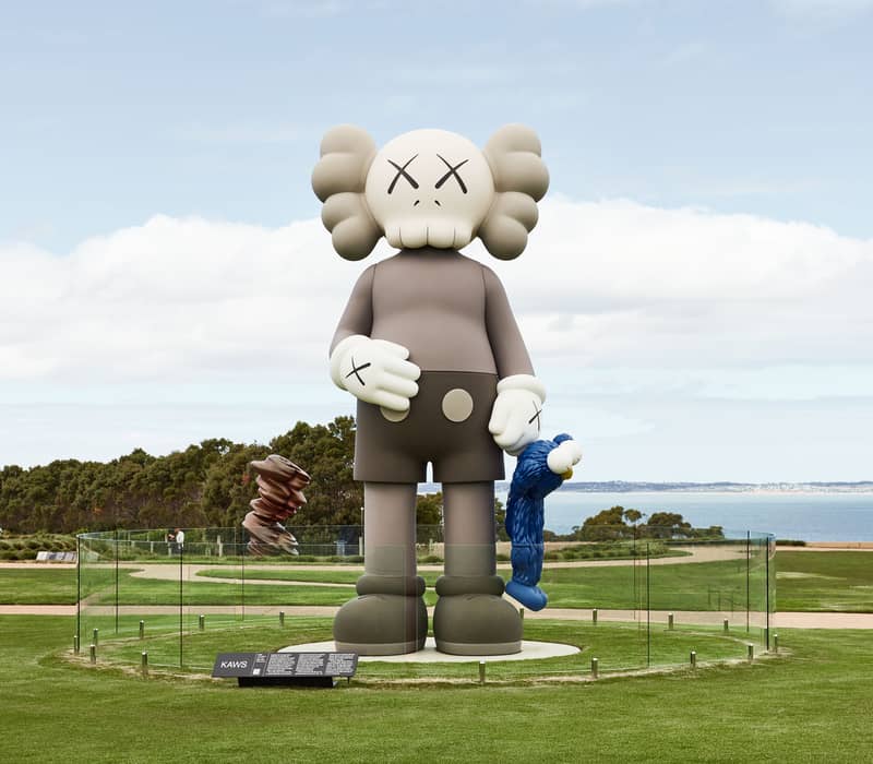 A large grey cartoon-like sculpture holding a small blue figure stands on a lawn with the sea behind it.
