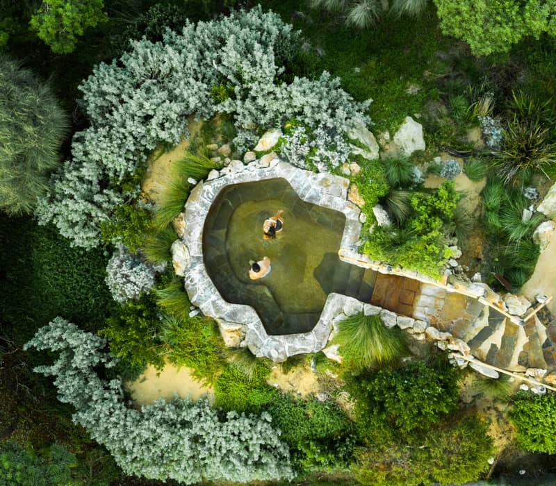 An overhead view of a natural stone pool nestled in green scrub with two people soaking in the water.