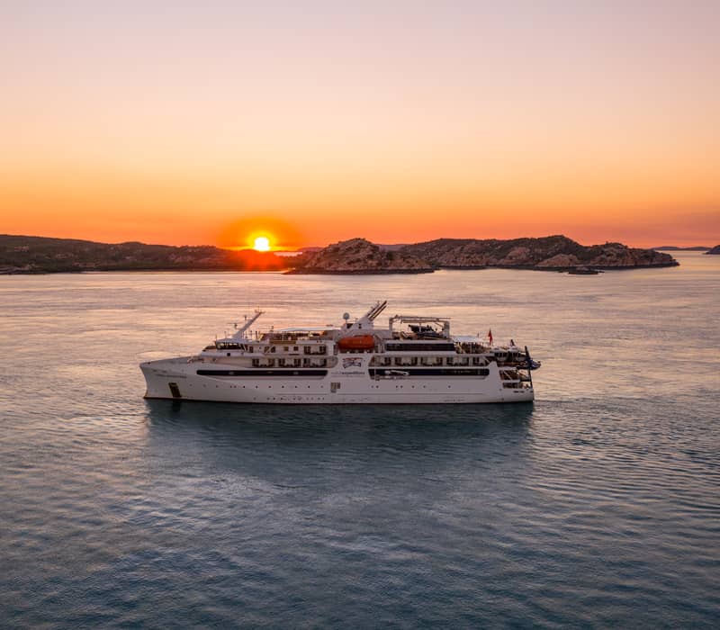 An APT luxury cruise ship positioned in the calm waters of the Buccaneer Archipelago during a vibrant sunset.