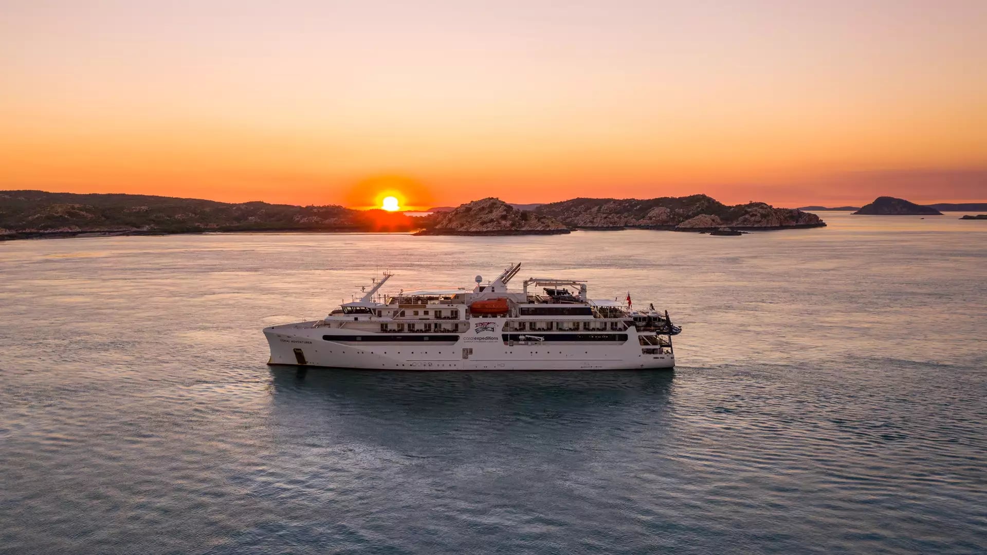 An APT luxury cruise ship positioned in the calm waters of the Buccaneer Archipelago during a vibrant sunset.