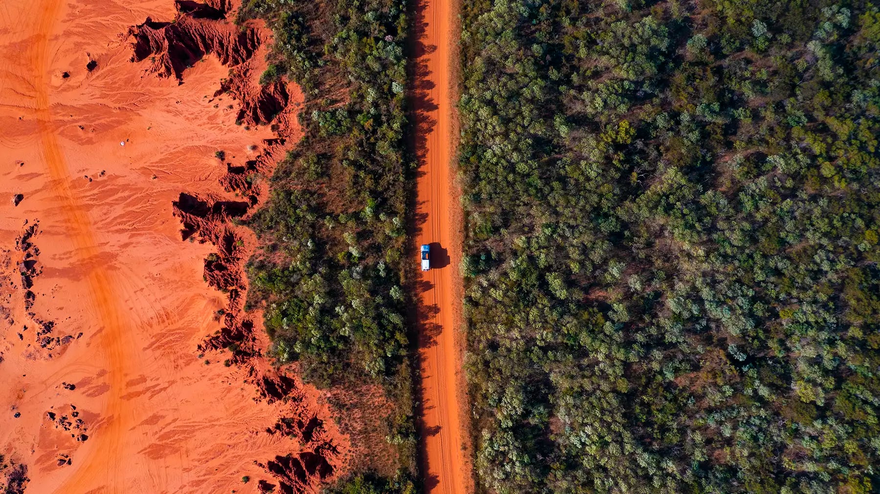 A vertical top-down aerial view of a straight red dirt road with a single white vehicle, flanked on one side by dense green bushes and on the other by eroded red earth formations.