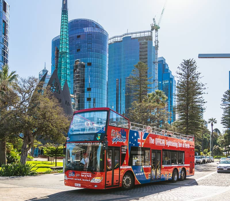 A red double-decker tour bus drives on a paved street in front of tall modern glass buildings in Perth.