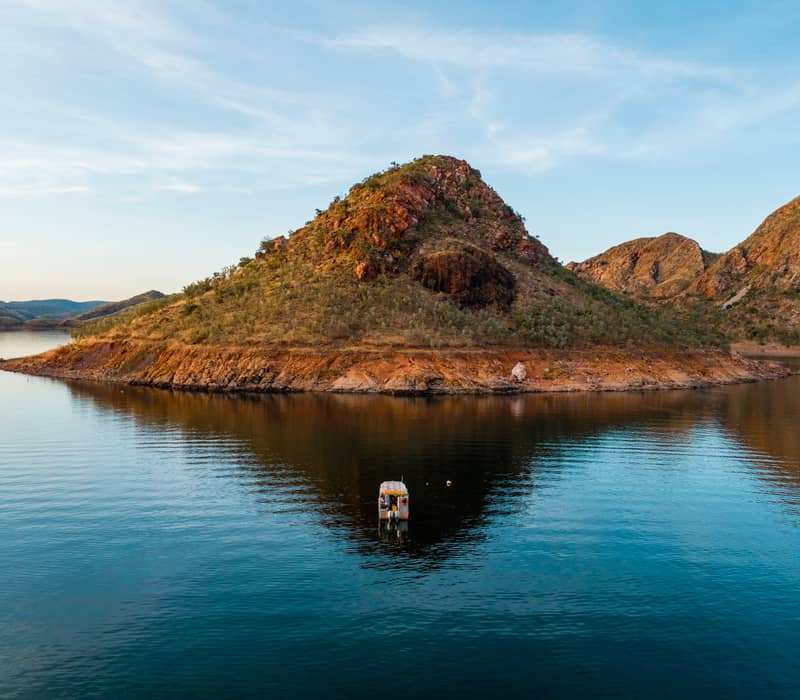 A small tour boat cruises on the calm blue water of Lake Argyle near a rugged red island in the Kimberley.
