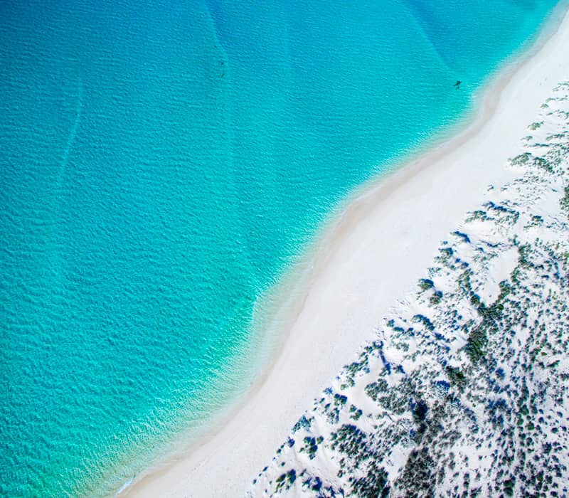An aerial view of a curved white sand beach meeting bright turquoise ocean water.