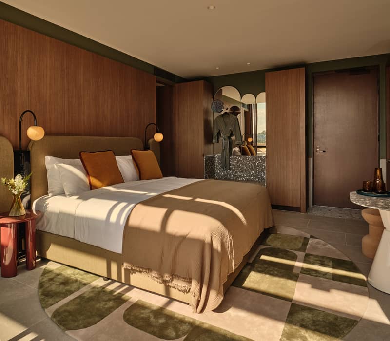 Elegant hotel suite interior with timber walls, a large bed, and sunlight streaming across a circular patterned rug.