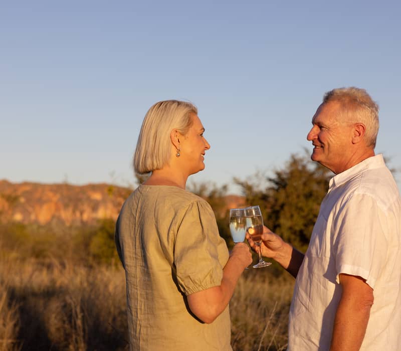 A man and woman clinking champagne glasses during sunset with orange mountains in the background.