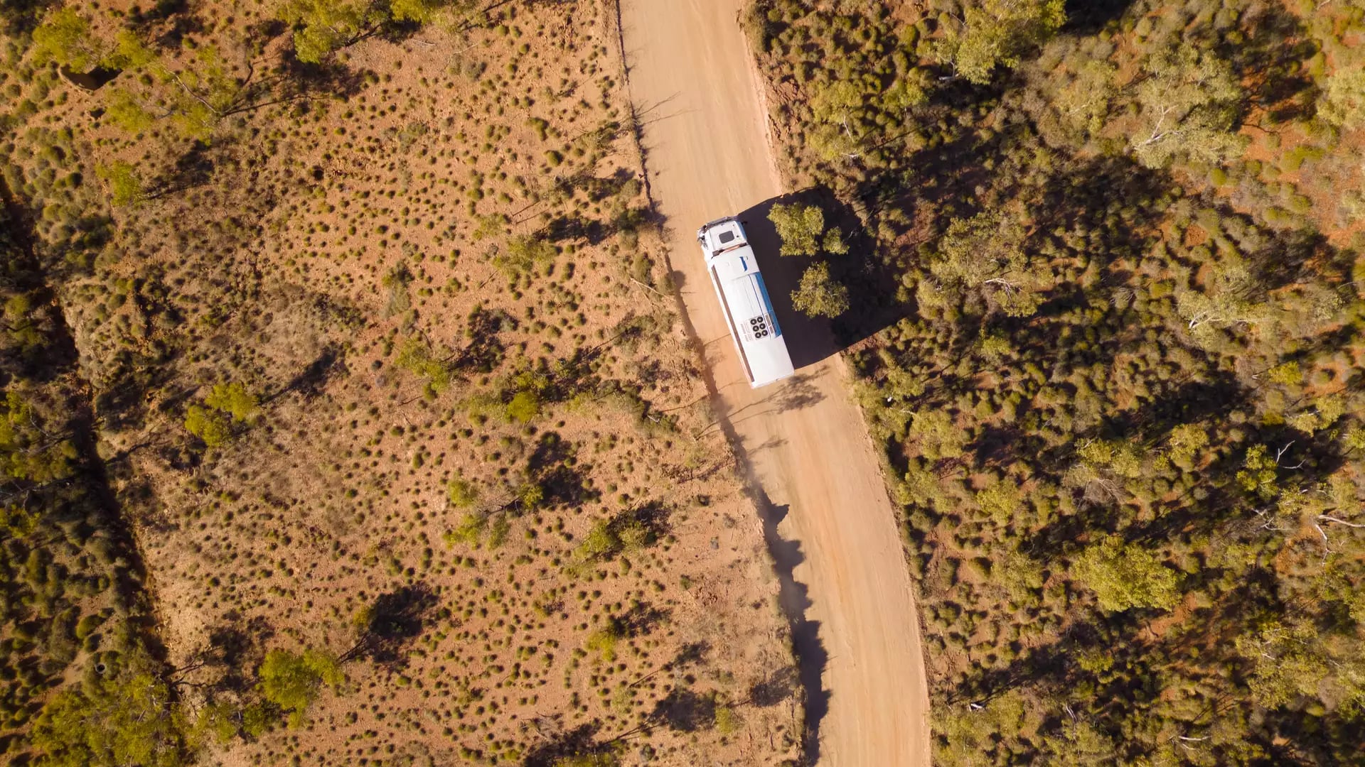 Top-down aerial shot of a white tour bus driving on a dusty orange outback road surrounded by green shrubs in Western Australia.