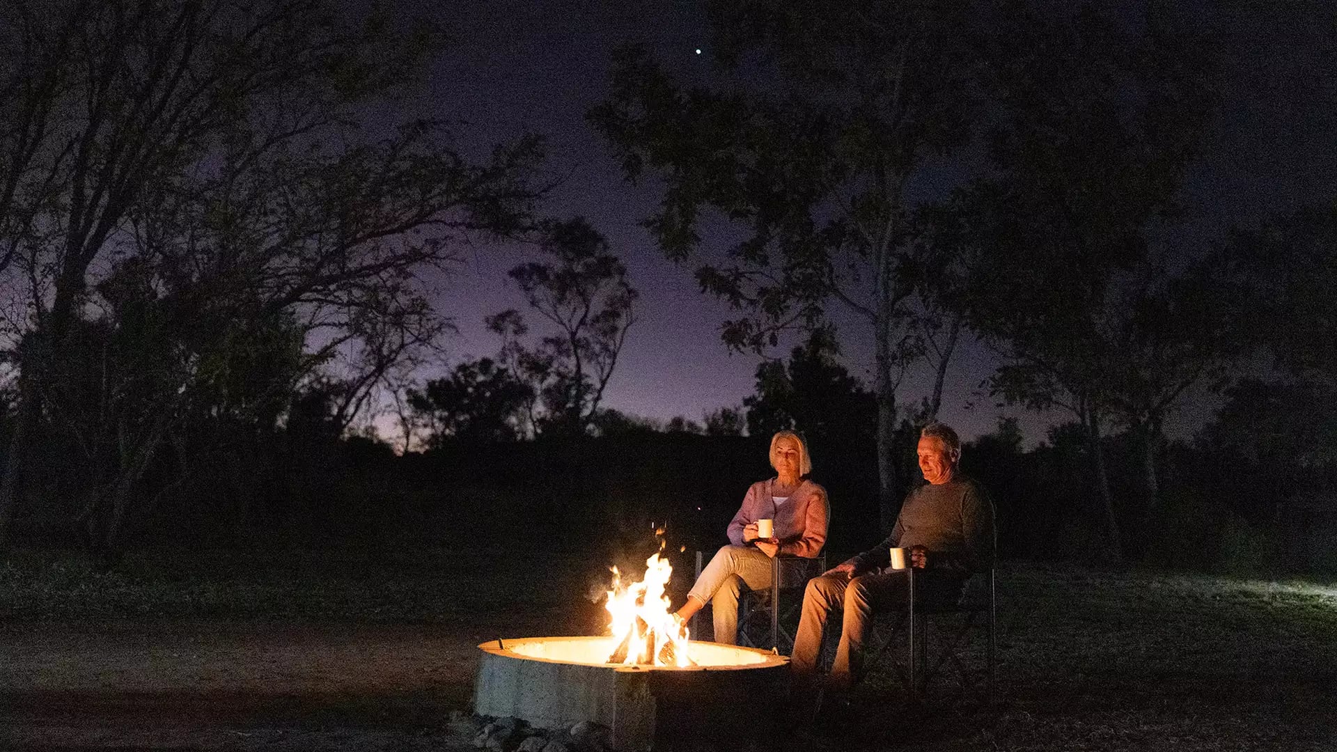 Two people sitting in chairs around a bright campfire at night under a purple dusk sky in the Kimberley.