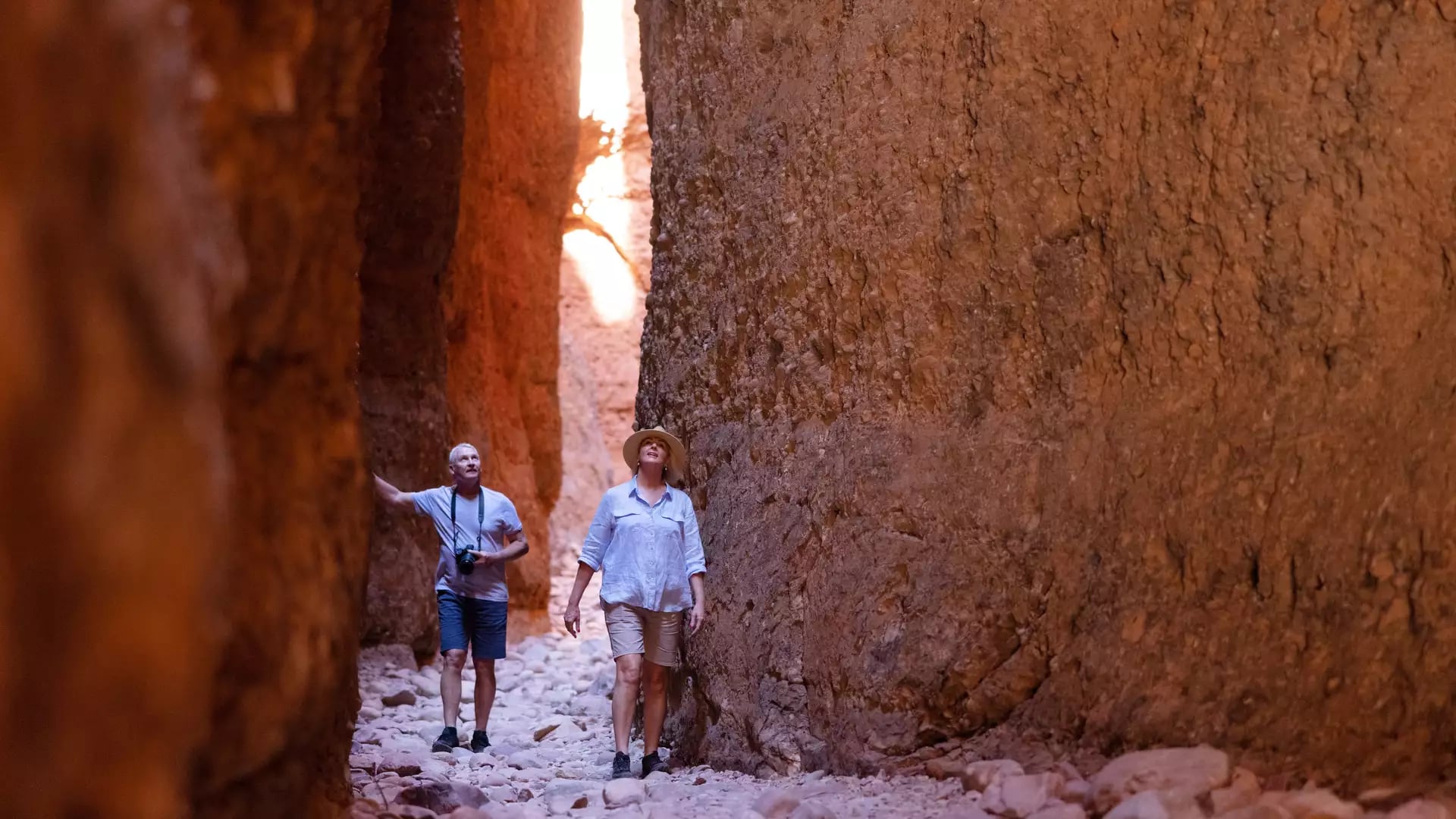 People walking on a rocky path through a very narrow and tall slot canyon with red stone walls.