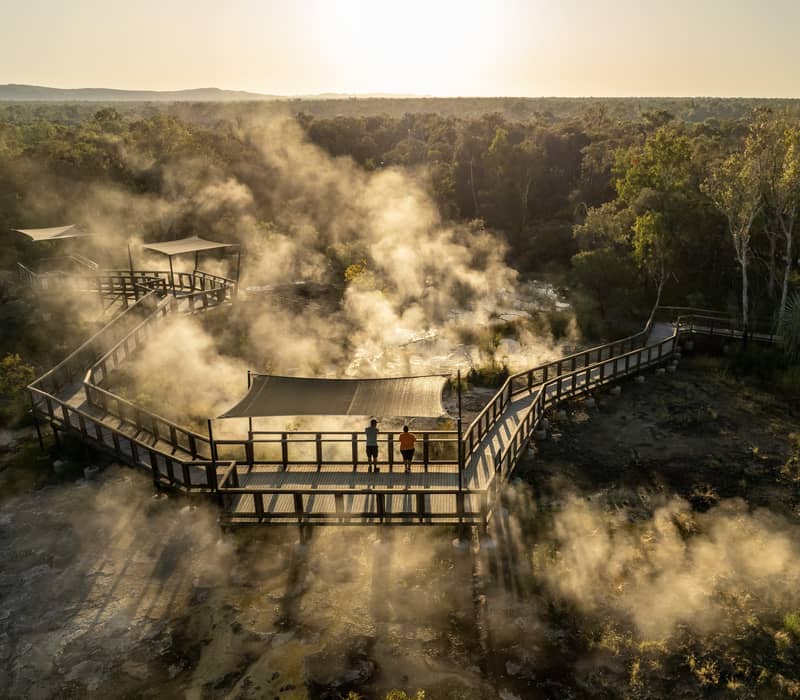 Aerial view of steam rising from hot springs with people on a wooden boardwalk at sunset in the outback.