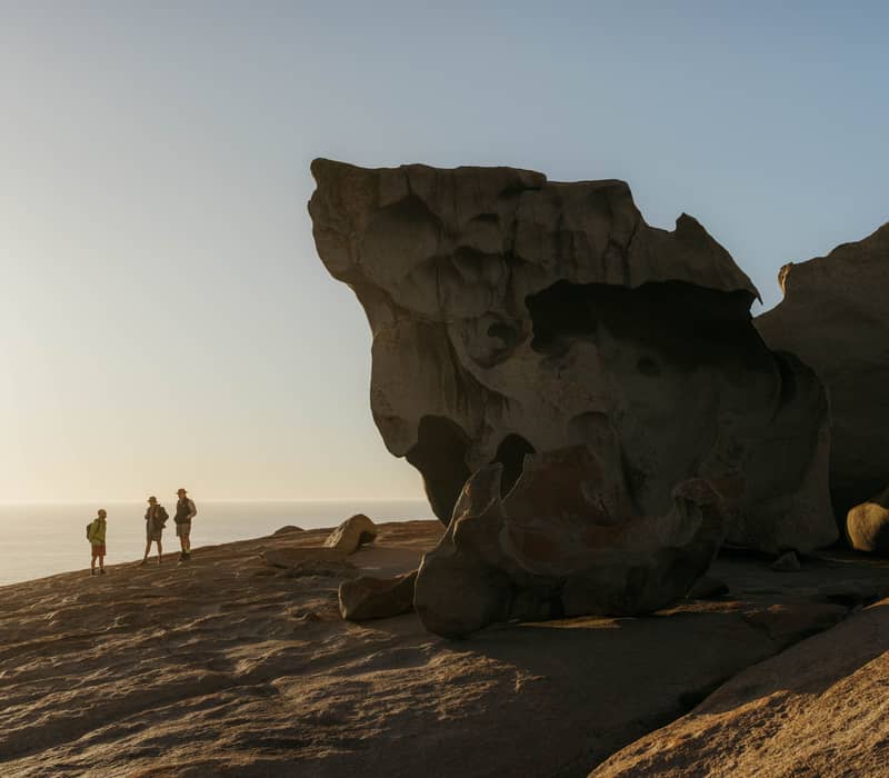 Three hikers standing next to the massive, weathered orange-lichen-covered granite boulders of the Remarkable Rocks against a pale blue sky.