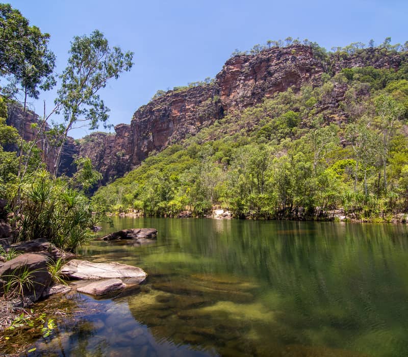 The crystal-clear, emerald-green plunge pool of Jim Jim Falls surrounded by steep sandstone cliffs and lush paperbark trees in the Mary River region of Kakadu.