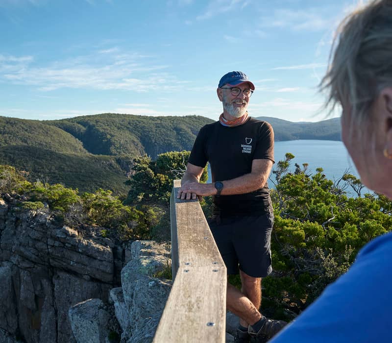 A male guide in a TWC uniform stands at a wooden railing overlooking a bay.