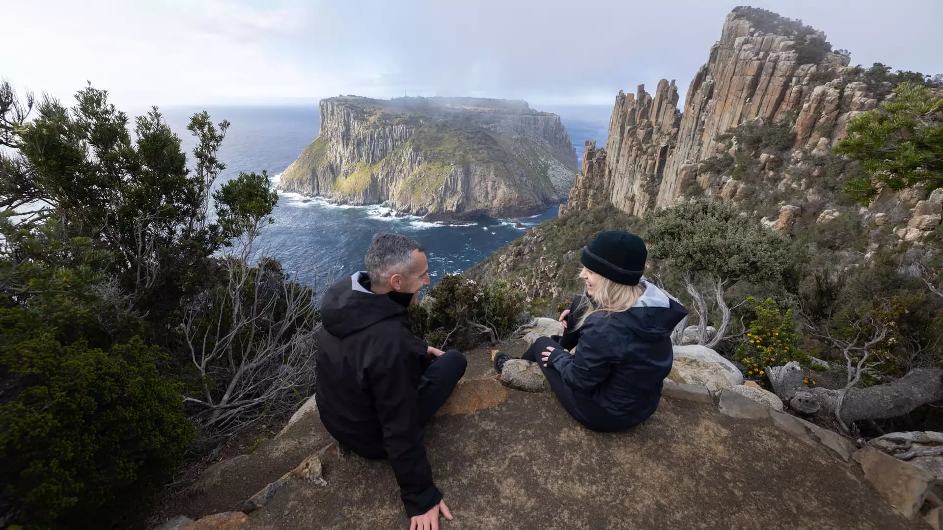 Two people sit on a rocky cliff edge with a view of a massive sea stack in the distance.