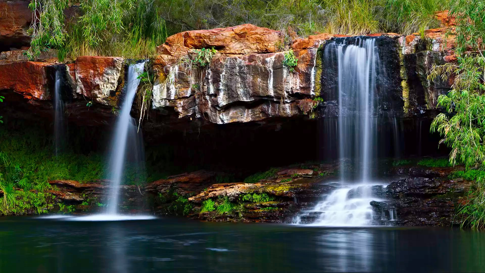 Twin waterfalls flowing over ancient red rocks into a dark green natural swimming pool surrounded by lush vegetation.
