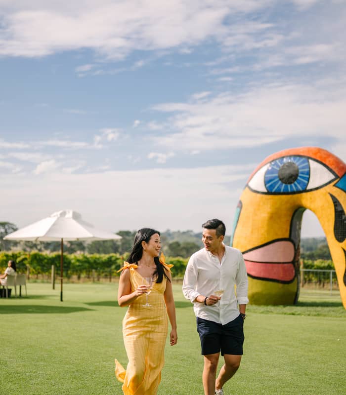 A man and woman walking on a green lawn past a large mosaic face sculpture at Pt. Leo Estate winery.