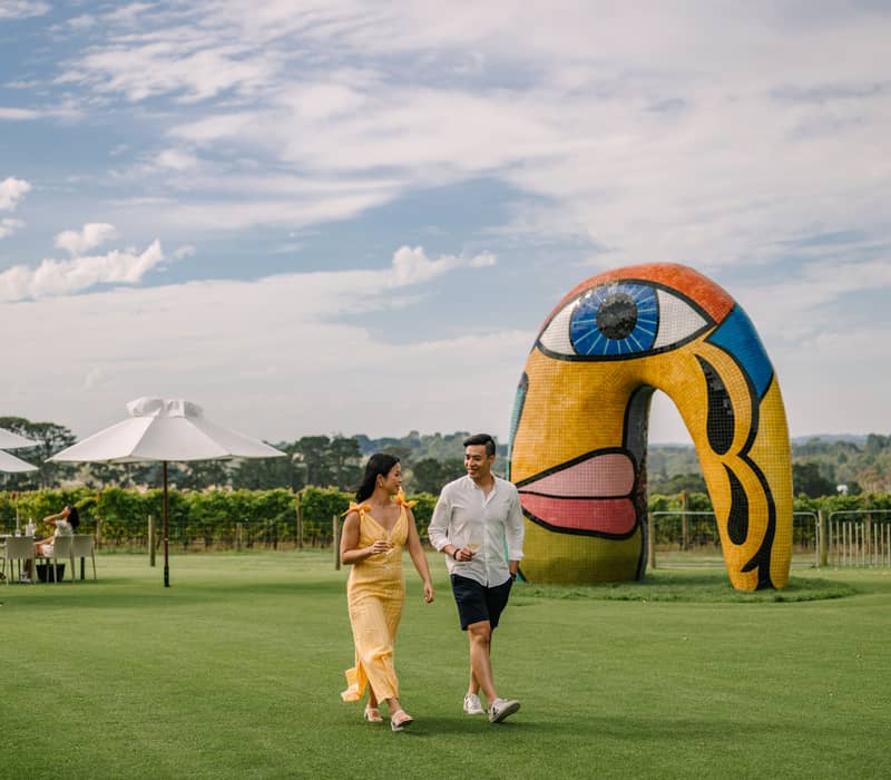 A man and woman walking on a green lawn past a large mosaic face sculpture at Pt. Leo Estate winery.