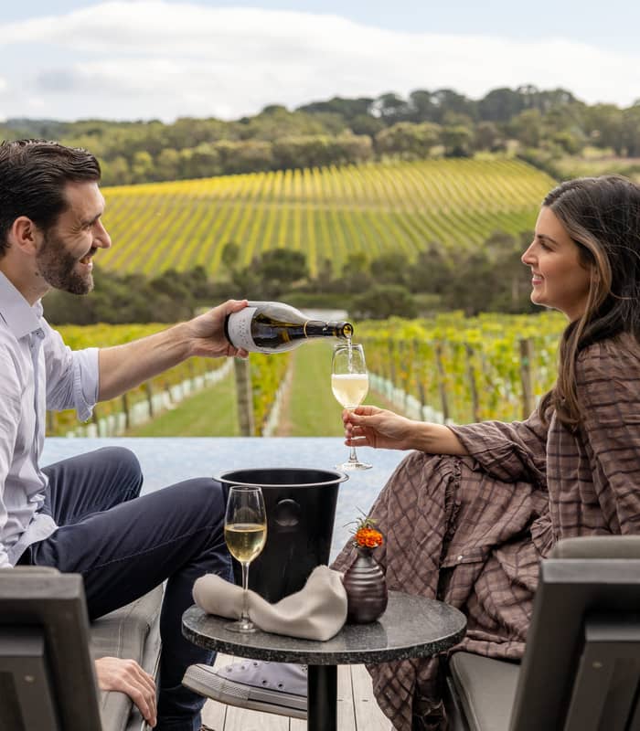 A man pouring wine for a woman sitting on lounge chairs next to an infinity pool overlooking a vineyard.