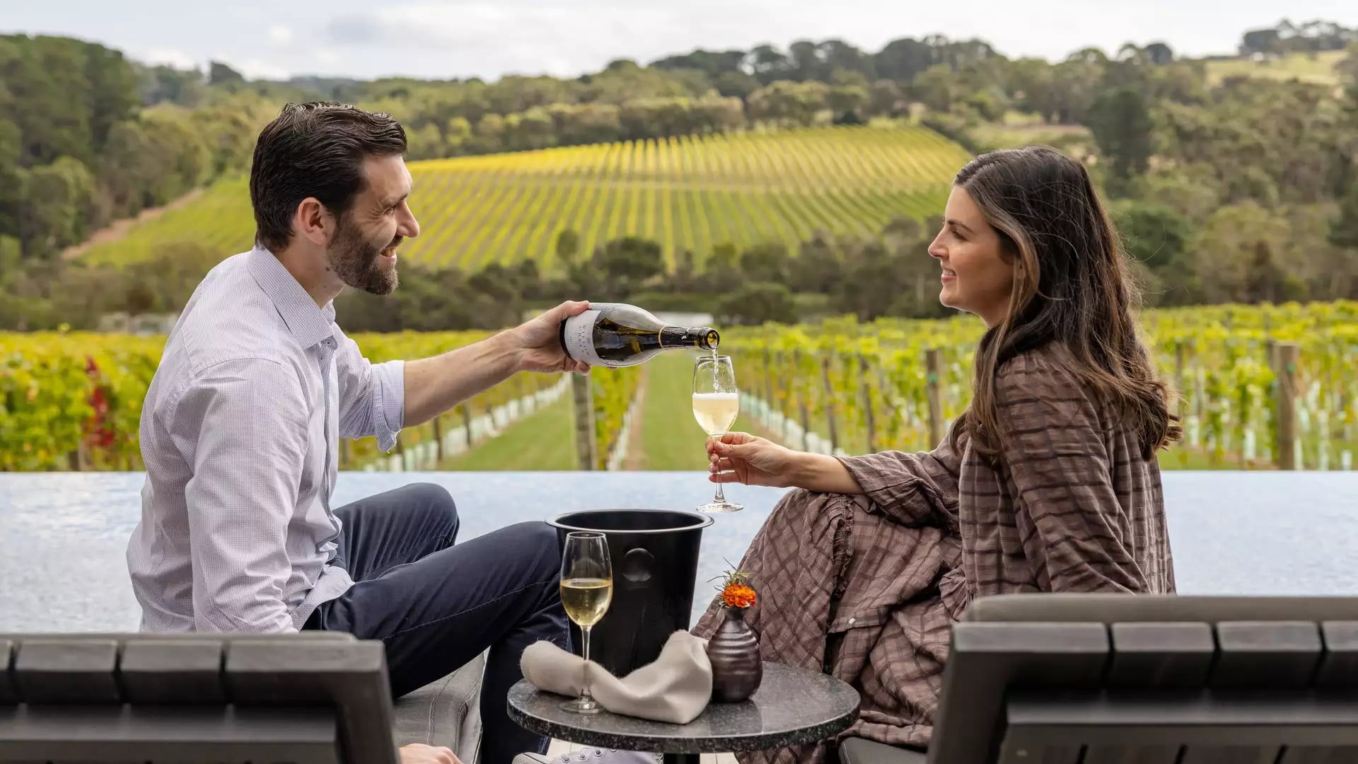 A man pouring wine for a woman sitting on lounge chairs next to an infinity pool overlooking a vineyard.